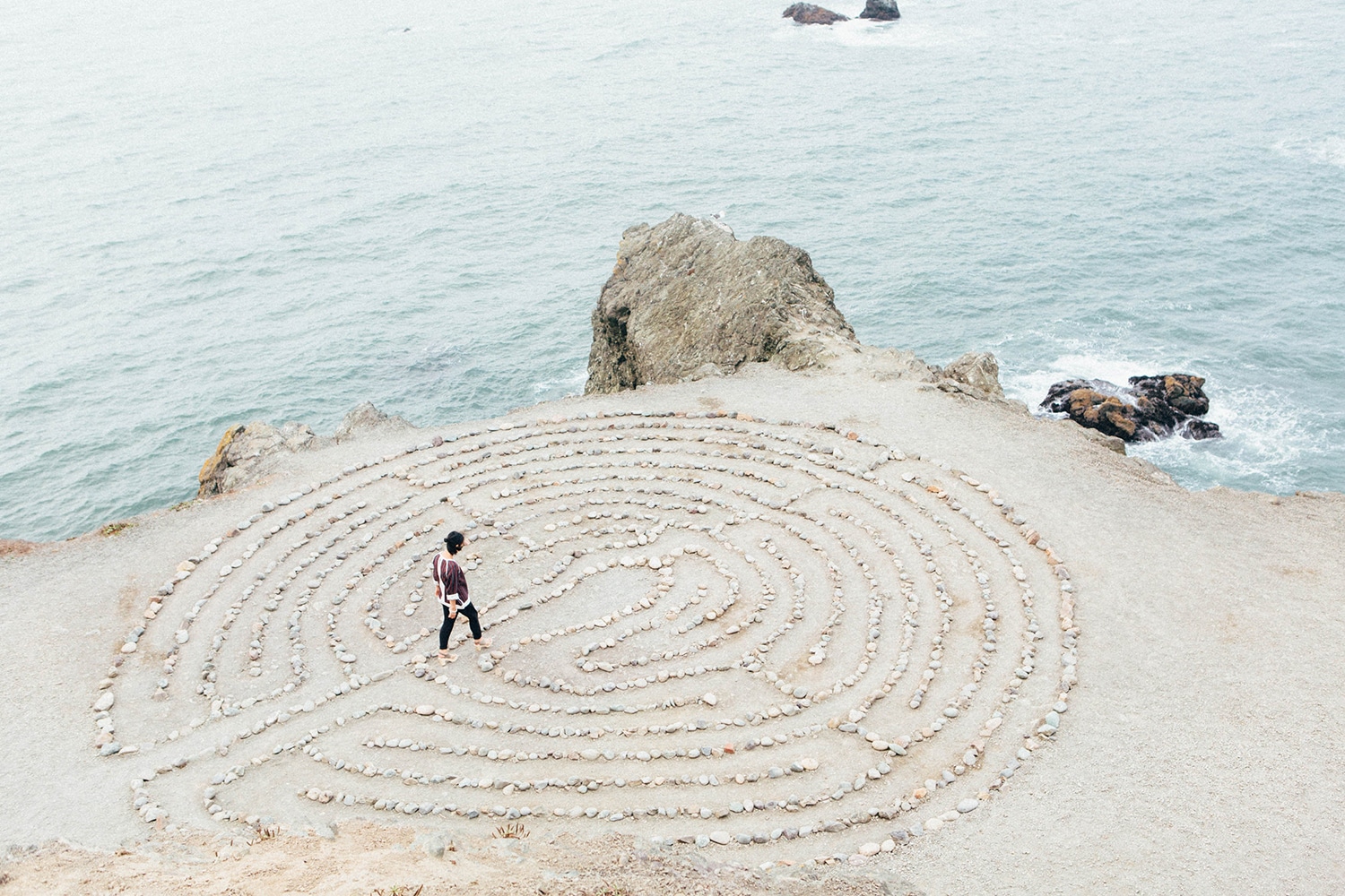 aerial view of person walking in large spiral sand drawing on top of a cliff next to water